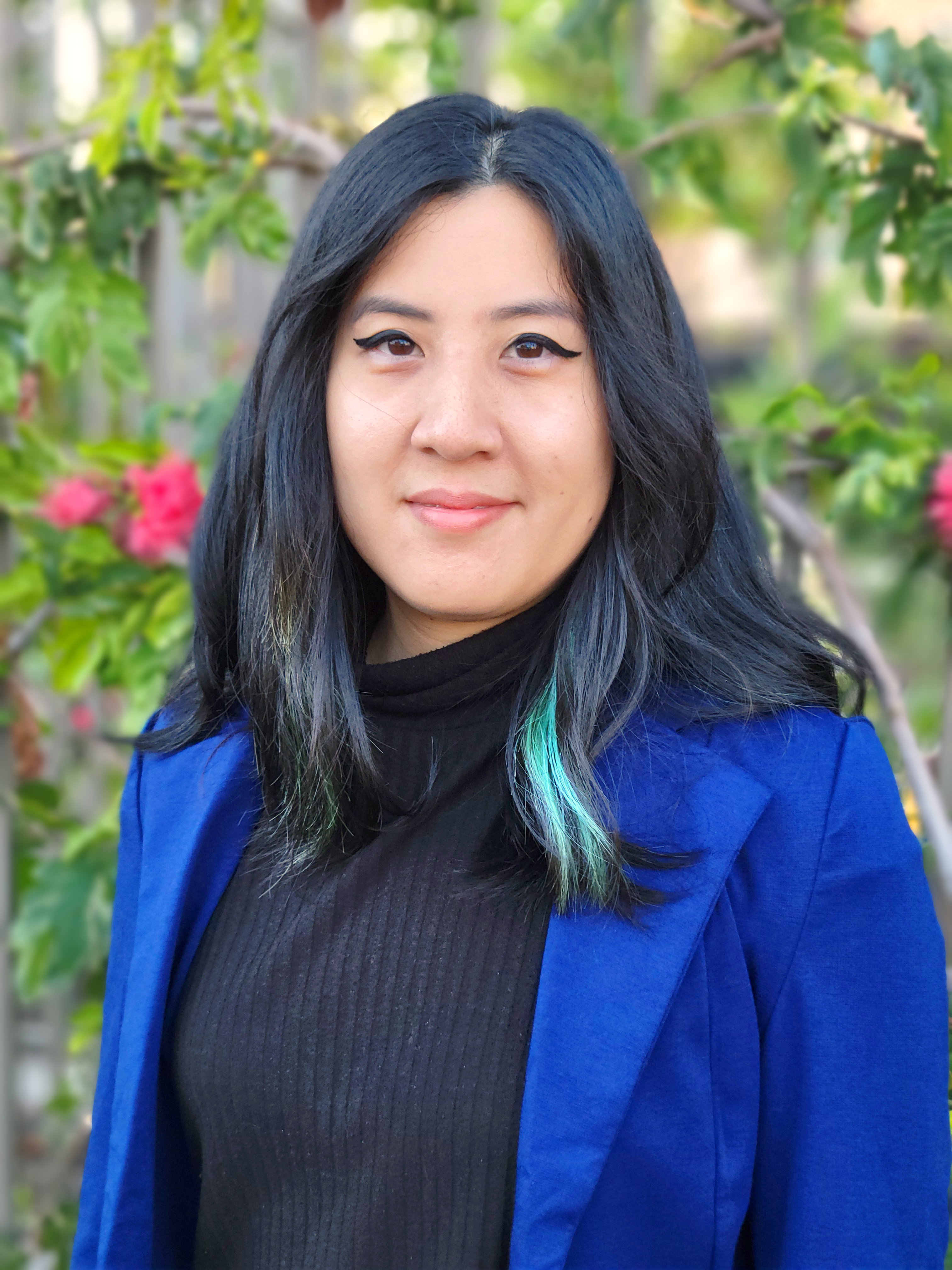 Thi, an Asian woman with black hair wearing black turtleneck and blue blazer with blurred foliage in background.